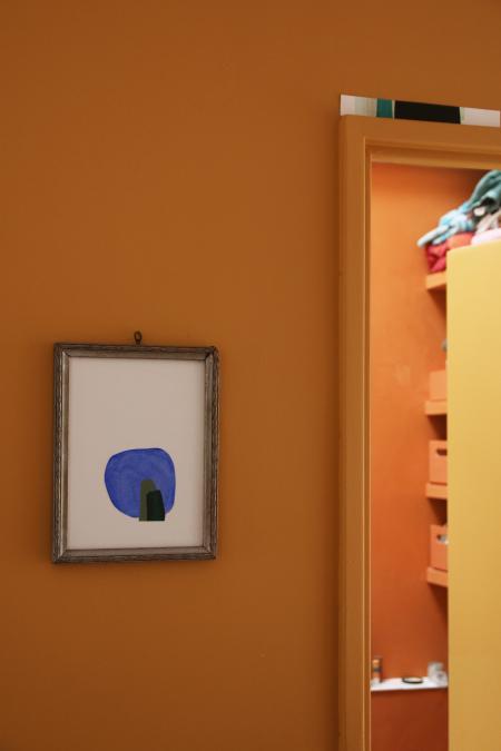 La porte entrouverte de la salle de bain sur mesure à Vertou imaginée par Jérôme Barrier donne à voir les étagères filantes entre 2 murs et des caisses de rangements, le tout peint en ocre et terracotta