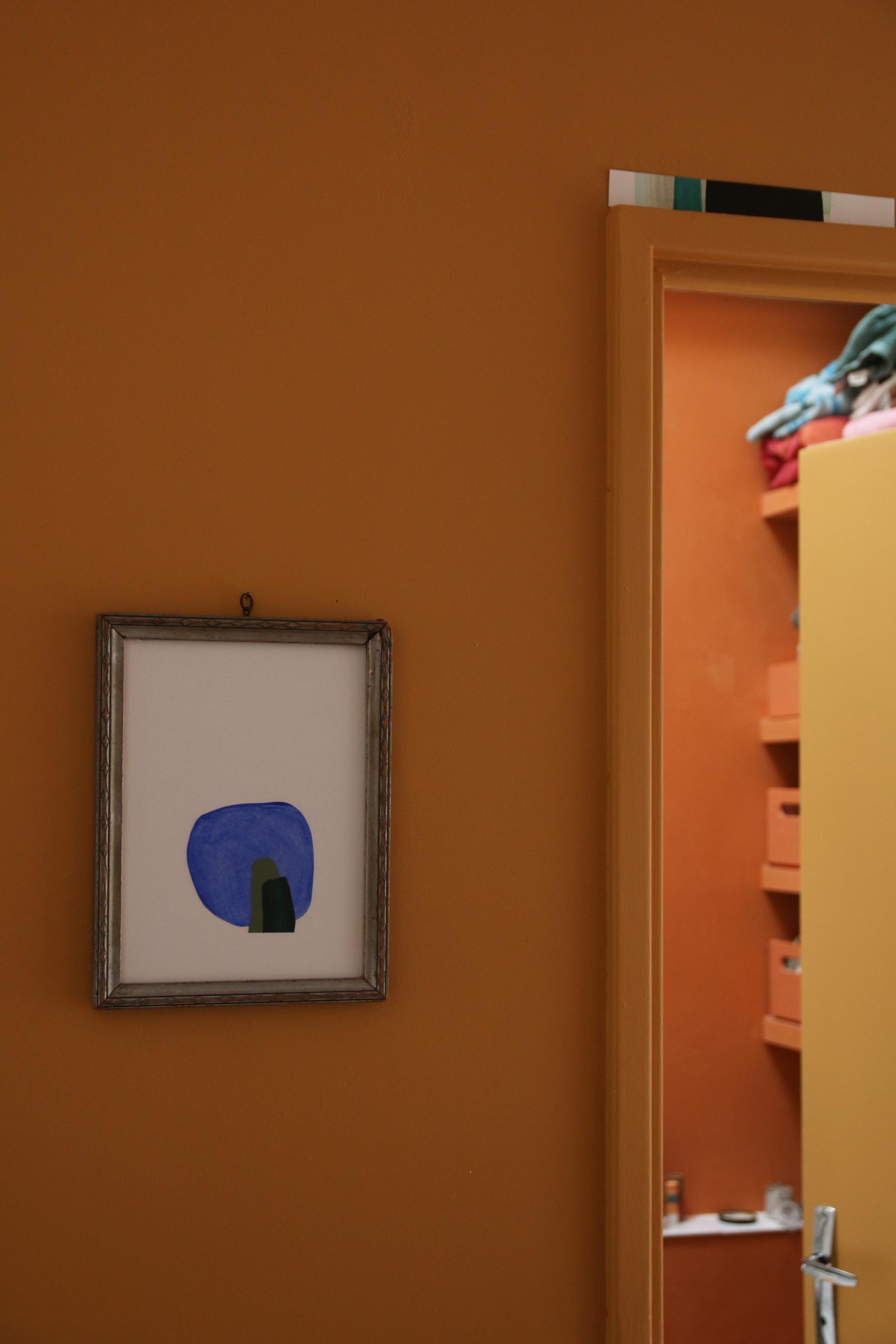 La porte entrouverte de la salle de bain sur mesure à Vertou imaginée par Jérôme Barrier donne à voir les étagères filantes entre 2 murs et des caisses de rangements, le tout peint en ocre et terracotta